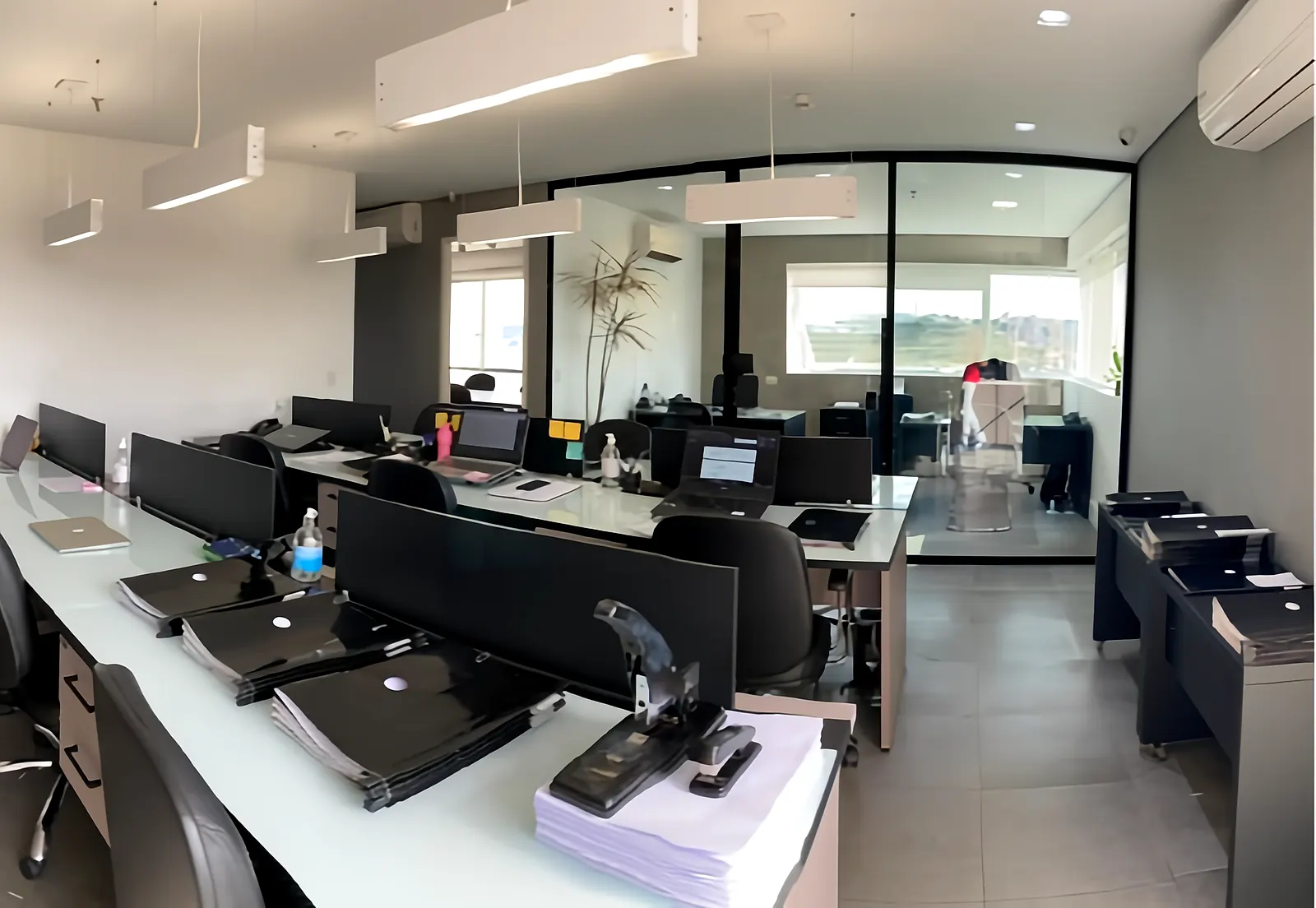 Área de trabalho da equipe do escritório, com estações e sala de reunião ao fundo